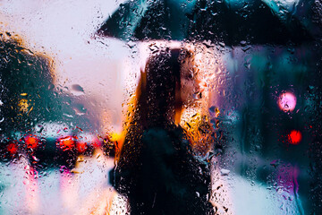 View through glass window with rain drops on blurred reflection silhouettesof a girl in walking on a rain under umbrellas and bokeh city lights, night street scene. Focus on raindrops on glass
