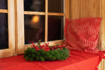 Fototapeta na wymiar Decorated Xmas wreath with candle and pillow on a windowsill. Comfort. Cozy Scandinavian decor of the room. Christmas Wreath of fir branches near window. Festive decoration at home 