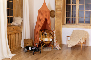 Rustic playroom interior with rattan crib with canopy, wicker baskets, toys and window. Wickered cradle bed with baldachin in newborn room in scandi style at home. Black racing car toy in a child room