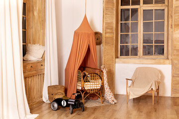Fototapeta na wymiar Rustic playroom interior with rattan crib with canopy, wicker baskets, toys and window. Wickered cradle bed with baldachin in newborn room in scandi style at home. Black racing car toy in a child room