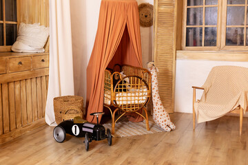 Rustic playroom interior with rattan crib with canopy, wicker baskets, toys and window. Wickered cradle bed with baldachin in newborn room in scandi style at home. Black racing car toy in a child room