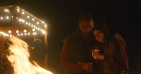 Multiethnic couple drinks tea, kisses and hugs while standing near the fire outdoors during...