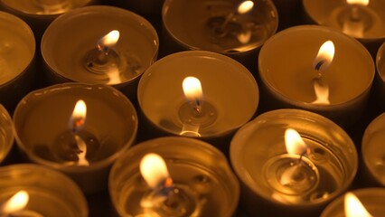 Many burning candles with melted wax. Soft warm light from the fire create a cozy atmosphere of peace and tranquility. Memorial day concept in church or temple, religion and ordinances. - obrazy, fototapety, plakaty