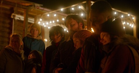 Group of multiethnic tourists talk while standing near the bonfire outside of wooden holiday cottage at night. Diverse friends or family resting outdoors during vacation in countryside or mountains.