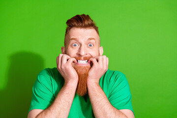 Portrait of horrified guy with red beard wear stylish t-shirt biting fingers astonished staring isolated on green color background - obrazy, fototapety, plakaty