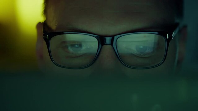 A laptop computer illuminates the face of a bespectacled programmer, who is intently analyzing the information on the screen. Information lines from the computer monitor are reflected in his eyes
