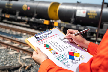 Action of an engineer is checking on chemical hazardous material checklist form with background of train freight tanker for crude oil or chemical cargo. Industrial safety working scene. 