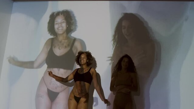 Horizontal medium long studio shot of young Black and Caucasian women in underwear dancing together with their image projected on backgroundHorizontal medium long studio shot of young Black and Caucas