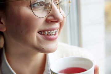 Close up portrait of young woman in dental braces and in glasses drinks hot tea while looking at window. Concept of medicine and beauty, health care and contemporary confidence.