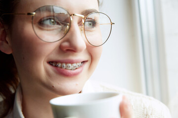 Close up portrait of young woman in dental braces and glasses drinks hot, warming coffee while...