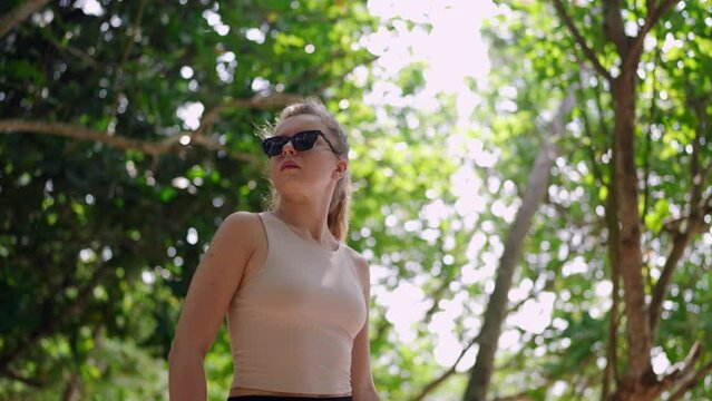Active woman in sunglasses enjoys nature walk in lush forest, breathing fresh air. Independent traveler female explores greenery, head held high. Solo adventure in serene trees, sunlight filtering.