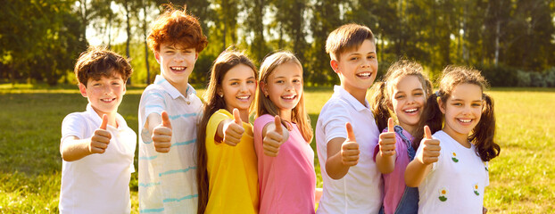 Cheerful kids enjoy summertime and spend free time on green meadow on good summer day. Banner header background with group of happy children standing in row showing thumbs up okay satisfaction gesture