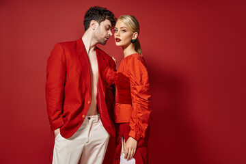 handsome man in red designer attire posing with blonde model on vibrant background, trendy couple