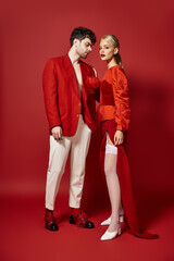 full length of handsome man in red designer attire posing with blonde model on vibrant background
