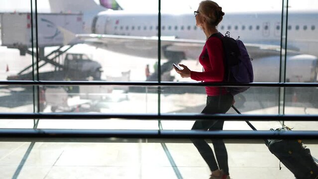 Profile view of happy attractive blond woman wearing backpack pulling luggage looking at phone walking with planes out of focus in the distance.