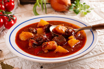 Pork goulash meat and potatoes on plate, garlic, onion, tablecloth in the background - typical Czech food