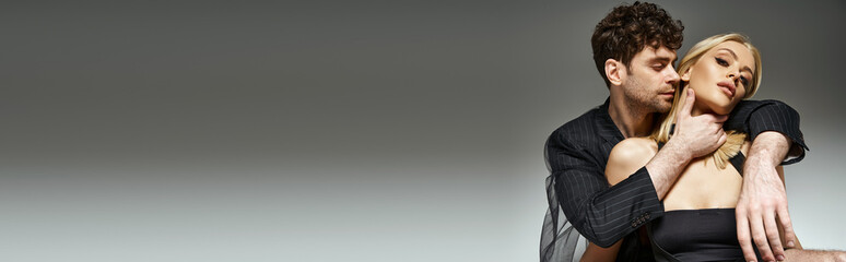 Fototapeta premium handsome curly man in pinstripe blazer embracing attractive blonde woman on grey background , banner
