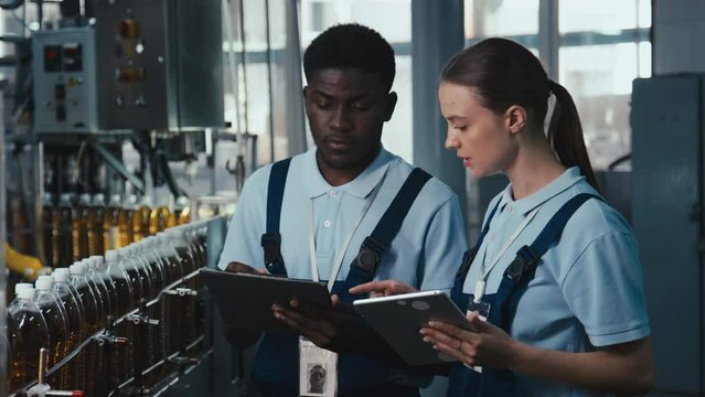 Medium footage of African American and Caucasian beverage plant specialists speaking about safety protocols standing with clipboard and tablet near conveyor with moving bottles of lemonade