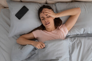 Young woman with headache lying in bed next to her phone