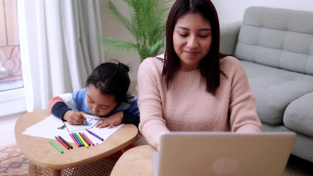 Latin American mother work on computer at home office. Child painting with business mom