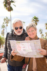 Naklejka premium Vertical elderly Caucasian tourist couple smiling holding travel map looking together interest places. Senior husband and wife standing in city enjoying pensioner vacation. Adult tourism people 