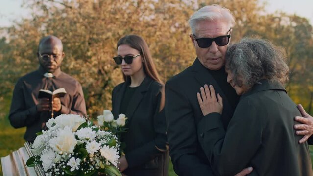 Medium footage of Caucasian senior man in sunglasses hugging his wife and comforting her during outdoor funeral service