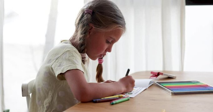Side view of preteen girl child in casual clothes looking down while sitting at table with various color pencils and writing notes in notepad near curtained window in daylight