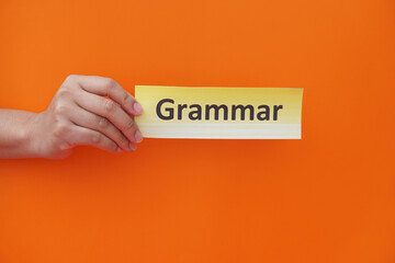 Close up hand holds paper card with text Grammar. Orange background. Concept, education, learning, studying language. Education. Reading and memorizing strategy of learning process. Grammar lesson.