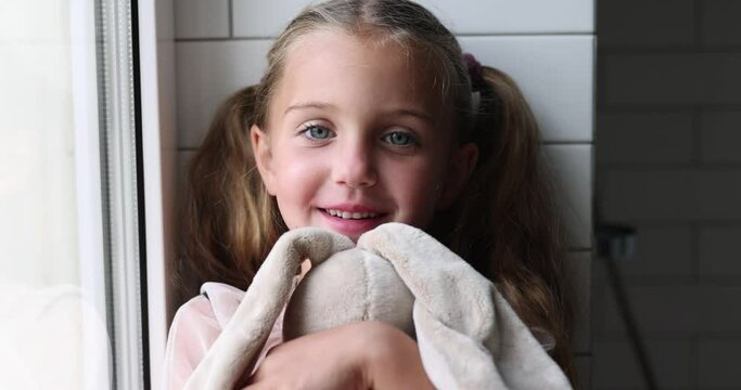 Cute smiling girl child with brown eyes and in ponytails looking at camera while sitting against tiles wall with folded legs in daylight and embracing stuffed rabbit toy in hands