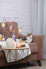Animal at home. Pet cat is lying on sofa in cosy living room. Ceramic vase with spring branches and easter eggs, burning candle on coffee table.