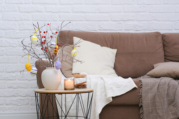 Vase with tree branches, Easter eggs and burning candle on table near the sofa in living room