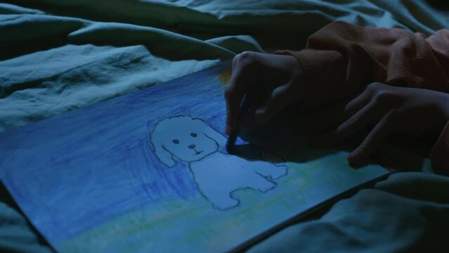 Closeup of hands of unrecognizable child drawing white puppy with colorful wax crayons in bed at night