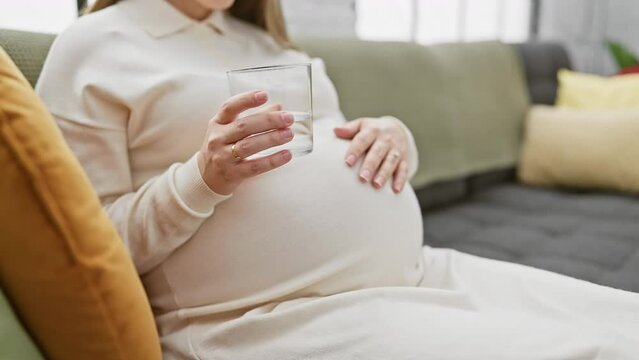A pregnant hispanic woman holding a glass sits on a couch indoors, depicting maternity and anticipation.