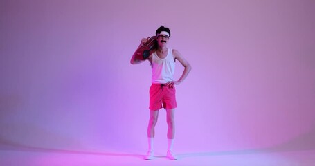 Friendly, funny young Caucasian 90s man with stereo player dancing, listening music, standing over purple background. Discotheque, party, entertainment concept.