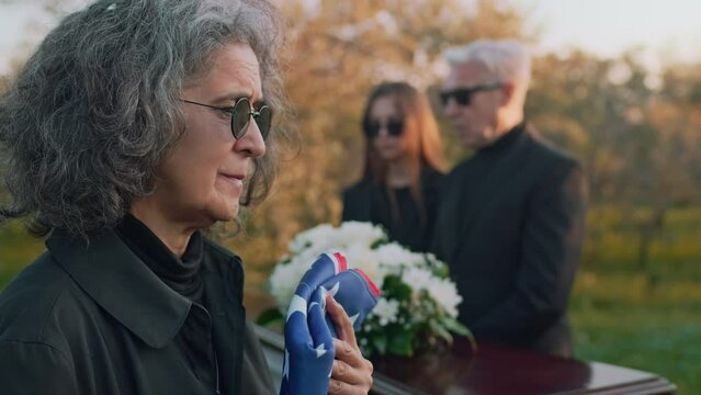 Side tilt closeup of unrecognizable man passing US flag to Caucasian mourning mother of deceased military soldier at outside funeral ceremony
