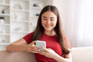 Smiling young asian woman focused on texting with her smartphone at home