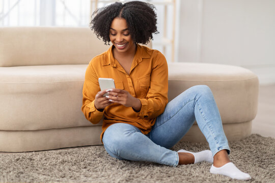 Joyful African American woman in a casual mustard shirt and blue jeans sits on the floor