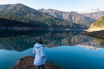 Woman standing at alpine lake Sauris (Lago di Sauris) in Friuli Venezia Giulia, Italy. Looking at water reflection of majestic mountain ridges of Carnic Alps. Serene tranquil atmosphere Italian Alps