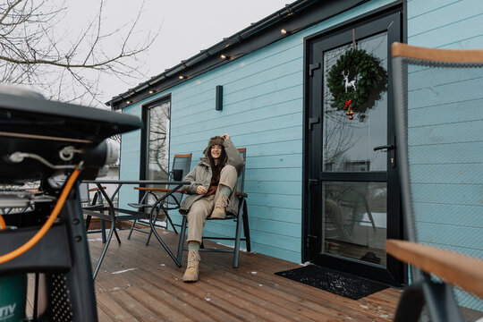 stylish girl in a warm jacket and hat in the yard of a tourist house is grilling meat and drinking coffee, enjoying a rural vacation and natural scenery