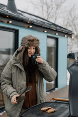 Countryside winter vacation for the weekend. Woman with a mug of warming drink against the background of a rented wooden house with a gas grill and Christmas decorations