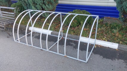 There is a metal structure on the asphalt sidewalk with a curb near a metal bench as bicycle parking. Ornamental bushes grow next to it near the metal profile fence