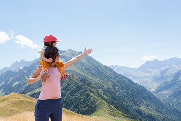 Smiling mother carrying her little daughter on shoulders with raised arm showing the mountain landscape.