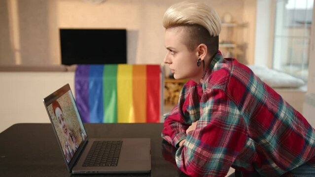 A woman in a plaid shirt and undercut hairstyle smiles while engaging in a video call, with a rainbow LGBT flag draped behind her, creating a warm atmosphere in the comfort of her home.