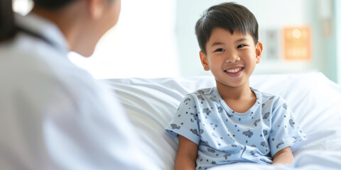 A young boy in a hospital gown smiles while sitting on a bed, facing an out-of-focus person, likely a medical professional, in a clinical room environment. Banner.