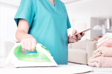 Caucasian woman, housewife, housekeeper, maid ironing clothes with an electric iron on an ironing board and looking at the screen of a mobile phone. Home interior background. soft focus.