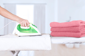 Caucasian woman, wife, mother, housewife, housekeeper, maid ironing bath towels in pink shades, electric iron on ironing board. Home interior background. soft focus.