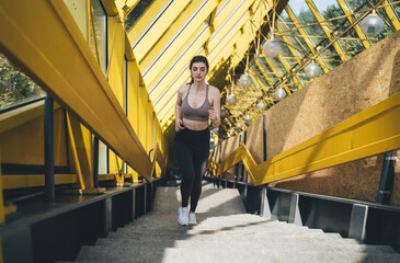 Fototapeta na wymiar Focused young Caucasian woman running down stairs in a vibrant yellow pedestrian overpass, showcasing fitness and urban active lifestyle in a sunny, dynamic environment
