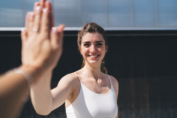 Radiant young Caucasian woman in a white tank top engaging in a high five, outdoors with a modern...