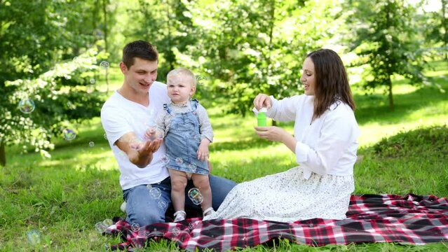 Happy caring parents and baby blowing soap bubbles while sitting on a blanket during a picnic in park.Young cheerful family is having a picnic in park.Summer time concept,outdoor recreation.Family day