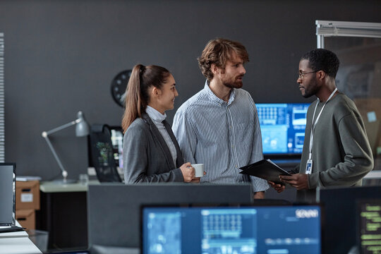 Over desktop view of three young people chatting standing in office with multiple computers copy space
